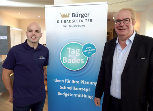 Marc Bürger vom Sanitärbetrieb Bürger Die Badgestalter und Jens J. Wischmann, Geschäftsführer der Vereinigung Deutsche Sanitärwirtschaft e. V. (VDS) lächeln in die Kamera, zwischen ihnen steht ein Banner mit dem Tag des Logos 2023 und dem Firmennamen und Logo des Betries Bürger Die Badgestalter,